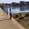 Moderner Hafenpromenade mit schwarzem Geländer, Kiesstrand und modernen Wohngebäuden am ruhigen Fluss